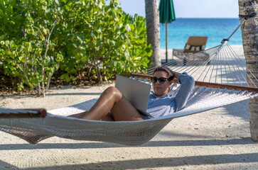 A young woman works on a laptop in a hammock on the beach in the Maldives. The concept of remote work and nomadism