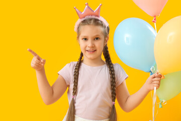 Little girl in crown with balloons pointing at something on yellow background. Children's Day celebration