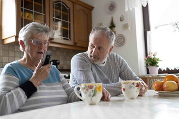 85 years old grey haired senior woman is holding a mobile telephone in the kitchen with her grey haired son 