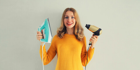Household chores, ironing - happy smiling woman housewife holding iron and hair dryer in her hands...