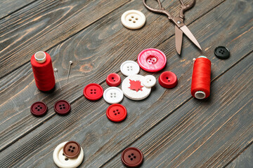 Buttons with maple leaf, thread spools and scissors on dark wooden background, closeup. Canadian flag concept