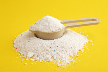 A pile of washing powder and a measuring spoon on a yellow background. Powder with blue and red...