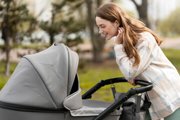 Smiling young mother smiling her baby, pushing stroller outdoors in park. Concept of care for child