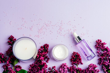 Aroma candle, cream jar and serum bottle with lilac flowers on light purple background. Natural cosmetics, skin care concept. Top view, flat lay, copy space