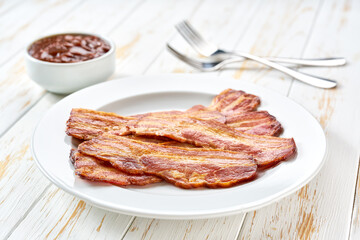 plate with fried bacon rashers and bbq sauce on a table, copy space for text. Plate with fried bacon on white kitchen table.