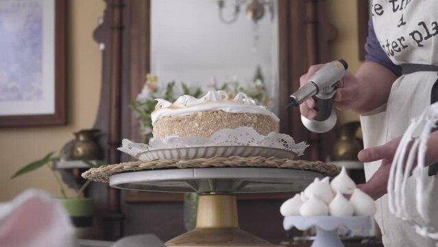 focus goes from meringues to pastry chef with torch burning sheet cake meringue icing on rotating base, home made environment