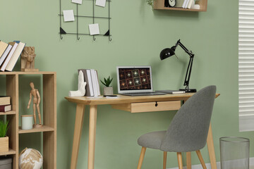 Workplace with modern laptop on wooden desk, bookcase and cosy chair at home