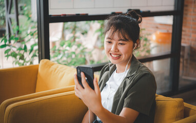 Smiling young asian woman using mobile phone checking social media,app playing game, shopping online, ordering delivery relax while sitting on a couch at cafe with laptop computer