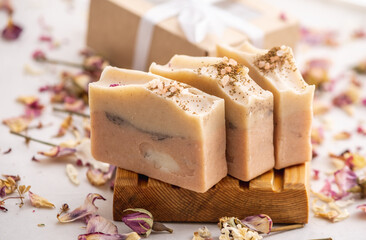 Pieces of delicate fragrant natural handmade soap on a wooden soap dish. On the table next to the soap is a gift box with a ribbon and dry flower petals