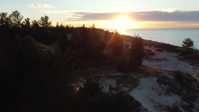 Aerial dolly over snowy forest landscape towards setting sun by water shoreline