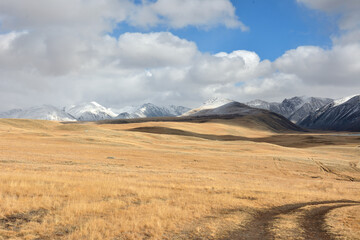 Fototapeta na wymiar Field roads cross high hills in the dry autumn steppe at the foot of high snow-capped mountains.