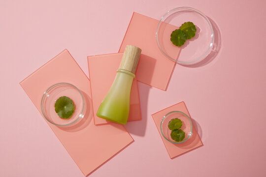 A green glass bottle placed on some acrylic sheets decorated on pink background with gotu kola leaves in petri dishes. Minimal scene for mockup packaging, cosmetics design.