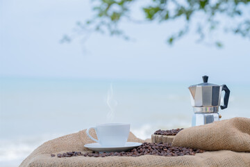 Close-up white coffee cup and many coffee beans placed around on the wood table with a beautiful seascape of nature background, concept coffee vacation travel.