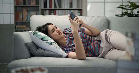 young asian female smiling and surfing the internet on a mobile phone while lying on the sofa