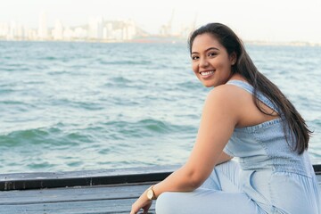 Latin woman is sitting on a pier, smiling on a summer day.