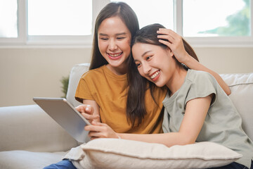 Fototapeta premium Happy lesbian, pleasure asian young two women, girl gay or close friend, couple love embrace, spending good time together, using tablet for entertainment on sofa at home. Activity of leisure, relax.