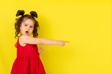 Smiling surprised little girl is attracted by attention pointing a finger at something. The concept of advertising goods and services. Isolated on yellow background