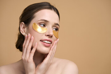 Young beautiful woman applying golden collagen patches under her eye, smiling over beige background