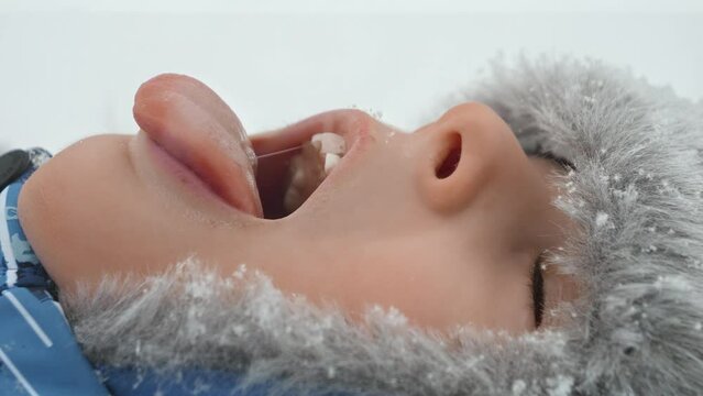 Portrait of boy lying in a snow-covered park, catching snowflakes with his tongue and smiling. Concept of outdoor winter play