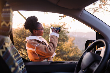 smiling black woman taking photos of nature with a camera during a road trip in camper van, concept of van life and travel adventures