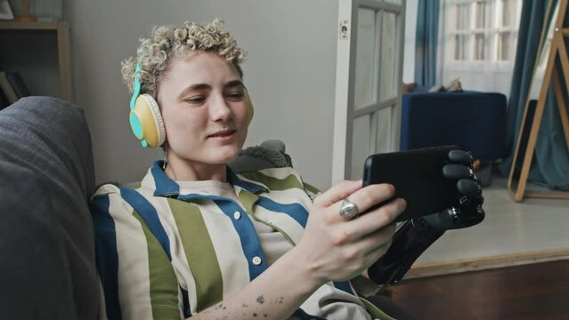 Stylish gen Z girl with short hair wearing arm prosthesis relaxing in living room listening to music in headphones and surfing Internet on smartphone