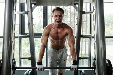 Naklejka premium Portrait of sweaty positive sportsman leaning on barbell and smiling at camera