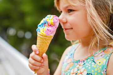 Happy preschool girl eating colorful ice cream in waffle cone on sunny summer day. Little toddler...