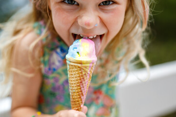 Happy preschool girl eating colorful ice cream in waffle cone on sunny summer day. Little toddler child eat icecream dessert. Sweet food on hot warm summertime days. Bright light, colorful ice-cream