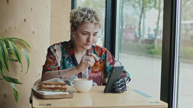 Stylish gen Z girl with short hair wearing prosthetic arm sitting at cafe table drinking cappuccino and surfing Internet on smartphone