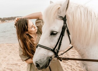 A woman in a dress stands next to a white horse on a beach, with the blue sky and sea in the...