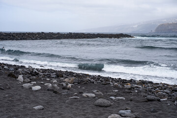 Black sand beach with waves. Breakwater on one side. Cliffs with houses in the background. Stones in the sand. Cloudy day. Puerto de la Cruz, Tenerife, Canary Islands, Spain.