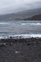 Black sand beach with waves. Breakwater on one side. Cliffs with houses in the background. Stones in the sand. Cloudy day. Puerto de la Cruz, Tenerife, Canary Islands, Spain.