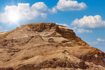 Fototapeta premium Background of mountains and sun in the blue sky in Dead Sea, Israel. Beautiful desert landscape among the sands and stones. Large stone ridge against the background of a blue sky with white clouds