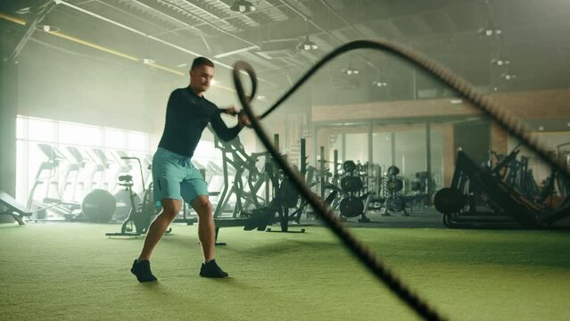 Grappler toss exercise on battle rope done by athletic young handsome man in gym with modern cardio area. Man has long sleeve compression shirt, shorts. High quality 4k footage Generative AI