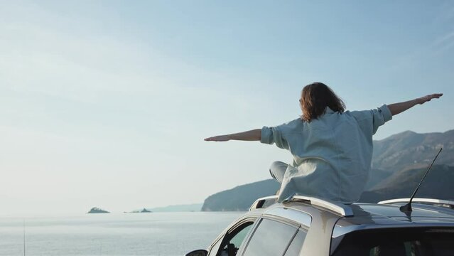 A young Caucasian woman sitting on the roof of her car parked by the sea and enjoying the beautiful view.