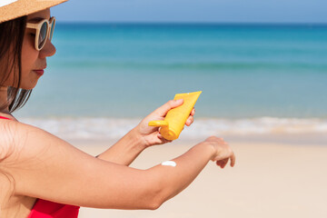 Asian woman wear a hat, sunglasses and red bikini apply sun cream on to her hand on tropical beach. Girl applying sunblock on her skin. UV protection, summer and beauty concept. Copy space, closeup
