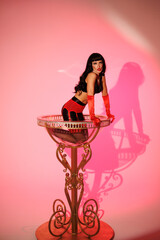 Pinup girl in a big glass like Dita Fon Tiz. Black wig, black underwear, stockings and red gloves, red light, red lipstick. Beautiful posch woman sitting in huge plastic goblet.