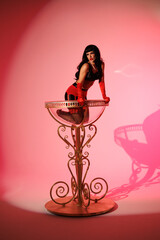 Pinup girl in a big glass like Dita Fon Tiz. Black wig, black underwear, stockings and red gloves, red light, red lipstick. Beautiful posch woman sitting in huge plastic goblet.