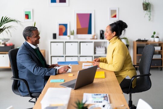 Side view of smiling Ethiopian woman and man in smart casual clothes talking while looking at each other in modern office