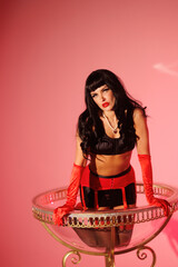 Pinup girl in a big glass like Dita Fon Tiz. Black wig, black underwear, stockings and red gloves, red light, red lipstick. Beautiful posch woman sitting in huge plastic goblet.