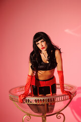 Pinup girl in a big glass like Dita Fon Tiz. Black wig, black underwear, stockings and red gloves, red light, red lipstick. Beautiful posch woman sitting in huge plastic goblet.