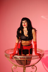 Pinup girl in a big glass like Dita Fon Tiz. Black wig, black underwear, stockings and red gloves, red light, red lipstick. Beautiful posch woman sitting in huge plastic goblet.