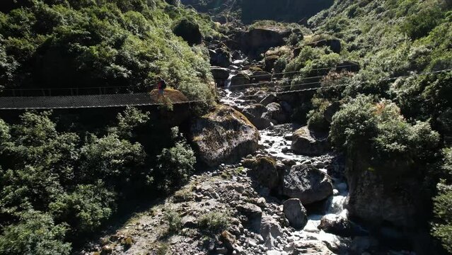 Young woman walking on swing bridge at Copland Track, New Zealand. Beautiful pristine native forest and river - drone reveal