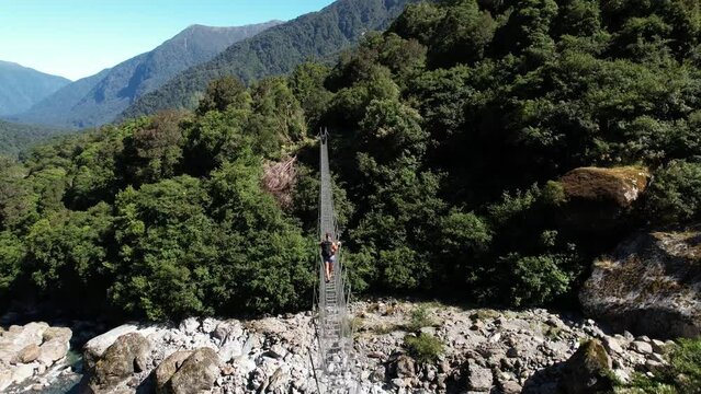 Aerial crane of girl on swing bridge crossing river in pristine nature. Copland track, New Zealand