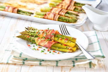 green asparagus wrapped in a roasted slices of bacon in a ceramic plate on a white wooden table
