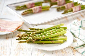 Prepared to bake appetizer, bundle of green asparagus wrapped in bacon on a baking tray on wooden table.