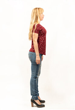 General shot of a beautiful blonde middle-aged girl facing the camera from right side in a photo studio, she is dressed casually in a red t-shirt, blue jeans and black shoes with big heels.