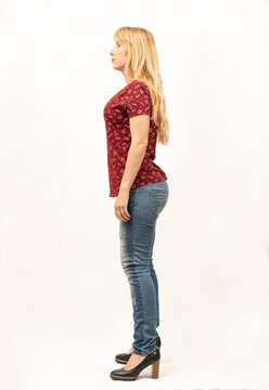 General shot of a beautiful blonde middle-aged woman facing the camera from the left side in a photo studio with white background, she is casually dressed in a red T-shirt, blue jeans and black shoes.