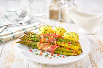 Homemade roasted bundles of green asparagus  wrapped in bacon on a ceramic plate on a white wooden table, close up.