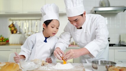 Asian father teaches child to break eggs onto the dough for baking cookies. Father and son in chef costume spends time together in kitchen at home